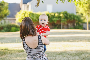 Wall Mural - Group portrait of Caucasian mother with baby boy in red t-shirt in field meadow outside. Female parent holding little happy smiling son child in summer park on sunset. Happy parenting lifestyle.