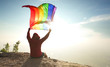 © 10incheslab - man sitting on mountain top raise rainbow LGBT symbol flag to bright sunny blue sky