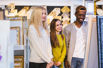 three multi ethnic people students reflecting at mirror with expensive frame indoor at frames store,