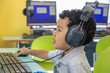 © Manny DaCunha - A small boy wearing oversized headphones navigates a wireless mouse in front of the computer. He remains focused on the computer.