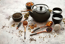 Iron Vintage Teapot And Cups. Dried Various Kinds Of Tea On Wooden Spoons. Stone Background With Copy Space