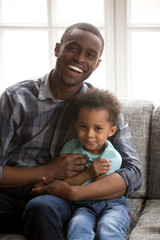 Wall Mural - African American family spend time together sitting looking at camera on couch in living room at home. Laughing father embrace shy smiling cute little preschool toddler son. Happy black family concept