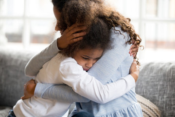 Black African mother embrace little preschool frustrated kid sitting on couch together at home. American loving mother supports disappointed daughter sympathizing, making peace after scolding concept