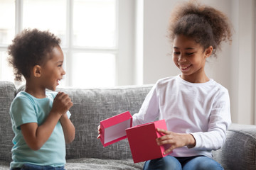 Black African attractive little siblings sitting on couch at home. Lovely toddler brother presenting giving pink gift box for sister. Boy congratulating a girl celebrating birthday life event concept