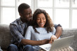 © fizkes - Cheerful black African married couple in love at home or hotel room. American wife husband hugging sitting on couch using computer looking on screen smiling feels happy spend time together on weekend
