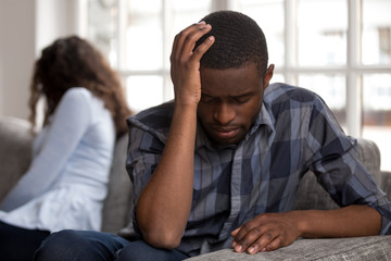 Close up black african couple sitting together not looking at each other on couch in living room at home after quarrel. Sad husband frustrated wife. Break up, problems trouble in relationship concept