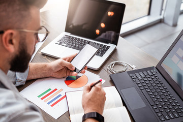 Wall Mural - Young man, entrepreneur, freelancer sits in office at table, uses smartphone, working on laptop, makes notes in notebook