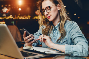 Young businesswoman is sitting in coffee shop at table in front of computer and notebook,using smartphone. Social media.