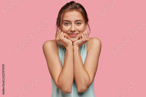 Fotografija Satisfied woman with delighted expression, keeps hand under chin, has healthy skin, green eyes, dark hair, wears casual clothes, isolated over pink background