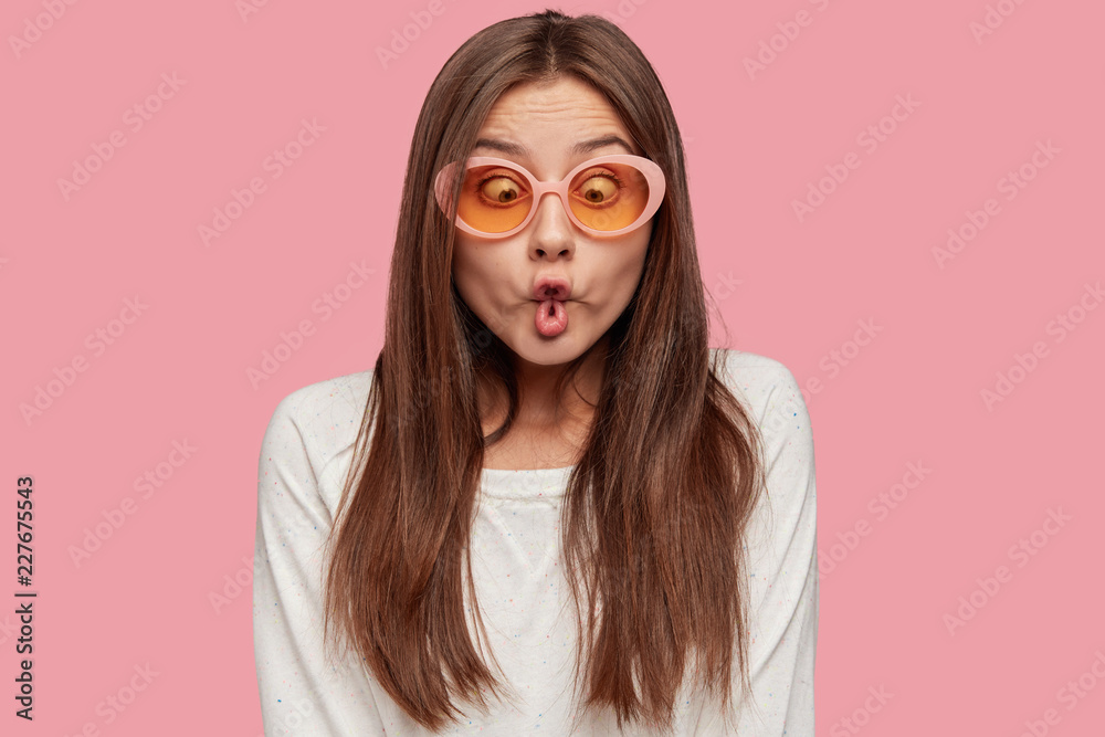 Funny playful Caucasian woman makes fish lips, has crazy look, tries to amuse depressed friend, feels bored, wears trendy shades and white sweater, isolated over pink background. Facial expressions - obrazy, fototapety, plakaty 