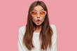 Funny playful Caucasian woman makes fish lips, has crazy look, tries to amuse depressed friend, feels bored, wears trendy shades and white sweater, isolated over pink background. Facial expressions