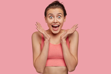 Isolated shot of overjoyed smiling young female fitness model in pink top clasps hands, has sporty body, feels amazed to find out about winning sport competition, isolated over pink background