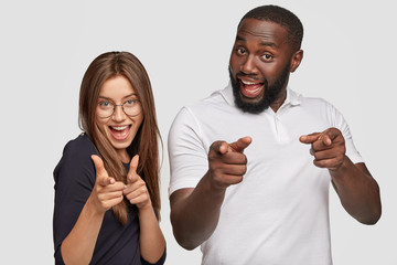 Positive girl and guy of different races make finger gun gesture, smile positively, express their choice, dressed in casual outfit, pose against white studio wall. We choose you. Bang, you are killed