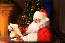 Portrait Of Happy Santa Claus Sitting At His Room At Home Near Christmas Tree And Reading Christmas Letter Or Wish List.