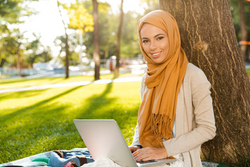 Poster - Photo of islamic female student 20s wearing headscarf, sitting on blanket in green park and using silver laptop