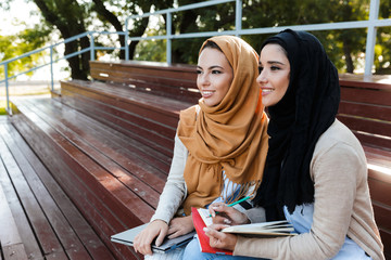 Canvas Print - Photo of joyous islamic girls wearing headscarfs, sitting on bench in park and studying