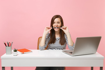 Passionate woman in casual clothes gnawing hold pencil in teeth sit work at white desk with contemporary pc laptop isolated on pastel pink background. Achievement business career concept. Copy space.