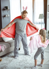 Wall Mural - I am super hero. Cheerful father is playing with his little daughter at home. He is wearing mantle and rabbit ears. Man is standing and laughing 