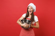 Housewife female chef cook or baker in striped apron white t-shirt toque chefs hat isolated on red wall background. Smiling surprised woman holding vacant crockery pot. Mock up copy space concept.