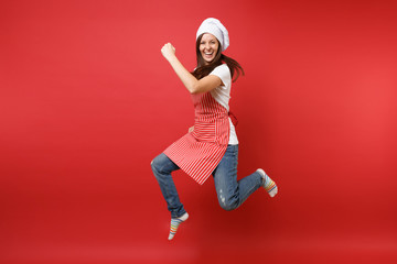 Housewife female chef cook or baker in striped apron white t-shirt, toque chefs hat isolated on red wall background. Full length portrait housekeeper woman jumping high up. Mock up copy space concept.