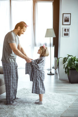 You are my heart. Happy father is holding hands with his little daughter. They are standing in living room and smiling. Family is wearing one pajamas on both and pink crowns 