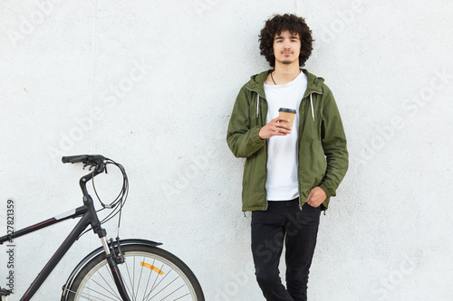 Fotografie, Obraz Indoor shot of handsome guy with curly hair, dressed in fashionable jacket, black trousers, drinks hot beverage, stands near bicycle against white concrete wall