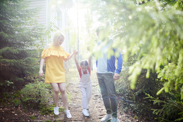 Wall Mural - Young blonde woman in yellow dress and her husband in casualwear holding their little daughter by hands while walking down forest path