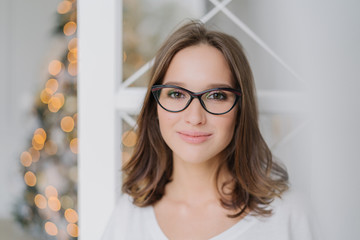 Close up shot of good looking young female model wears spectacles for good vision, looks happily at camera, has pleased expression, charming smile, poses against decorated Christmas tree indoor