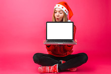 Excited By The Good News, The Woman In Santa's Hat, Holding A Blank Laptop Screen And Looking At It From Above, Copies The Space For Text, On A Red Background.