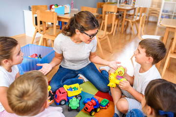 Wall Mural - Preschool teacher talking to group of children sitting on a floor at kindergarten