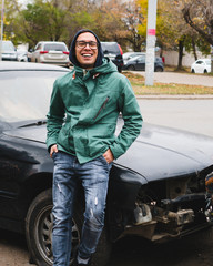 Man in glasses and jacket is smiling at the wrecked car