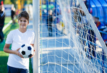 cute young boy in white blue sportswear holds classical black and white football ball on the green grass of the stadium field. Soccer game, training, hobby concept. with copy space 