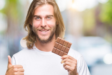 Young handsome man with long hair eating chocolate bar over isolated background happy with big smile doing ok sign, thumb up with fingers, excellent sign