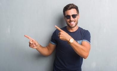 Handsome young man over grey grunge wall wearing sunglasses smiling and looking at the camera pointing with two hands and fingers to the side.