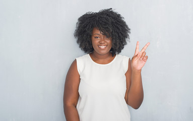 Sticker - Young african american woman over grey grunge wall smiling with happy face winking at the camera doing victory sign. Number two.