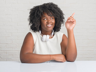 Poster - Young african american woman sitting on the table wearing headphones with a big smile on face, pointing with hand and finger to the side looking at the camera.