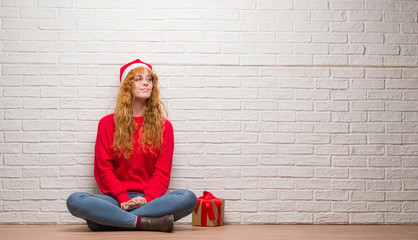 Poster - Young redhead woman sitting over brick wall wearing christmas hat smiling looking side and staring away thinking.