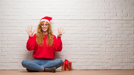 Wall Mural - Young redhead woman sitting over brick wall wearing christmas hat showing and pointing up with fingers number nine while smiling confident and happy.