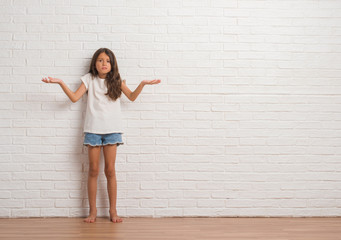 Poster - Young hispanic kid stading over white brick wall clueless and confused expression with arms and hands raised. Doubt concept.