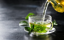 Hot Chinese Green Tea With Mint, With Splash Pouring From The Kettle Into The Cup, Steam Rises, Dark Background, Selective Focus