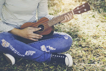 Asian teen girl wearing jeans and T-shirt playing ukulele is within the park. She was sitting on the lawn in the warm sunlight shines from the back.