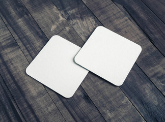 Photo of two blank white square beer coasters on vintage wooden background.