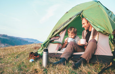 father with son drink hot tea sitting in camp tent