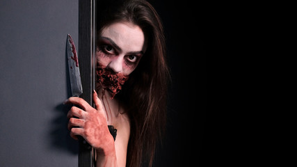 Makeup for halloween party. A girl with a bloodied knife in her hand looks out from behind a gray wall. Imitation of a bloody wound and a wired mouth. Black background. Copy space.