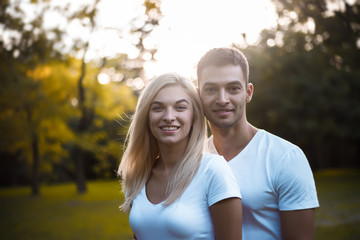 Wall Mural - Cute loving couple standing on grass in nature green park with beautiful sundown light. Looking camera.