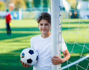 cute young boy in white blue sportswear holds classical black and white football ball on the stadium field. Soccer game, training, hobby concept. 