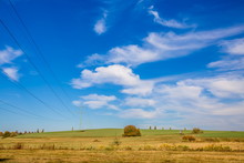 Autumn Landscape On A Bright Sunny Day, Blue Sky, White Clouds, Green And Brown Grass, Electricity Pylon With Wires Standing On Field, Horizon In Distance