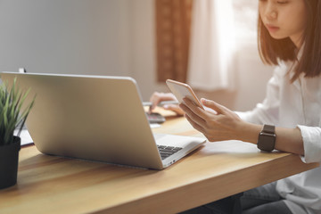 Fototapete - woman holding smartphone and using laptop on table in office room on windows with trees and nature background, for graphics display montage. Take your screen to put on advertising.