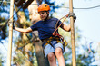 © Natali - Child in forest adventure park. Kid in orange helmet  and blue t shirt climbs on high rope trail. Agility skills and climbing outdoor amusement center for children. young boy plays outdoors.