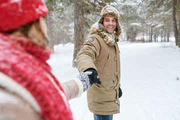 Wall Mural - Waist up portrait of handsome young man looking at girlfriend and smiling happily while leading her through beautiful winter park, copy space